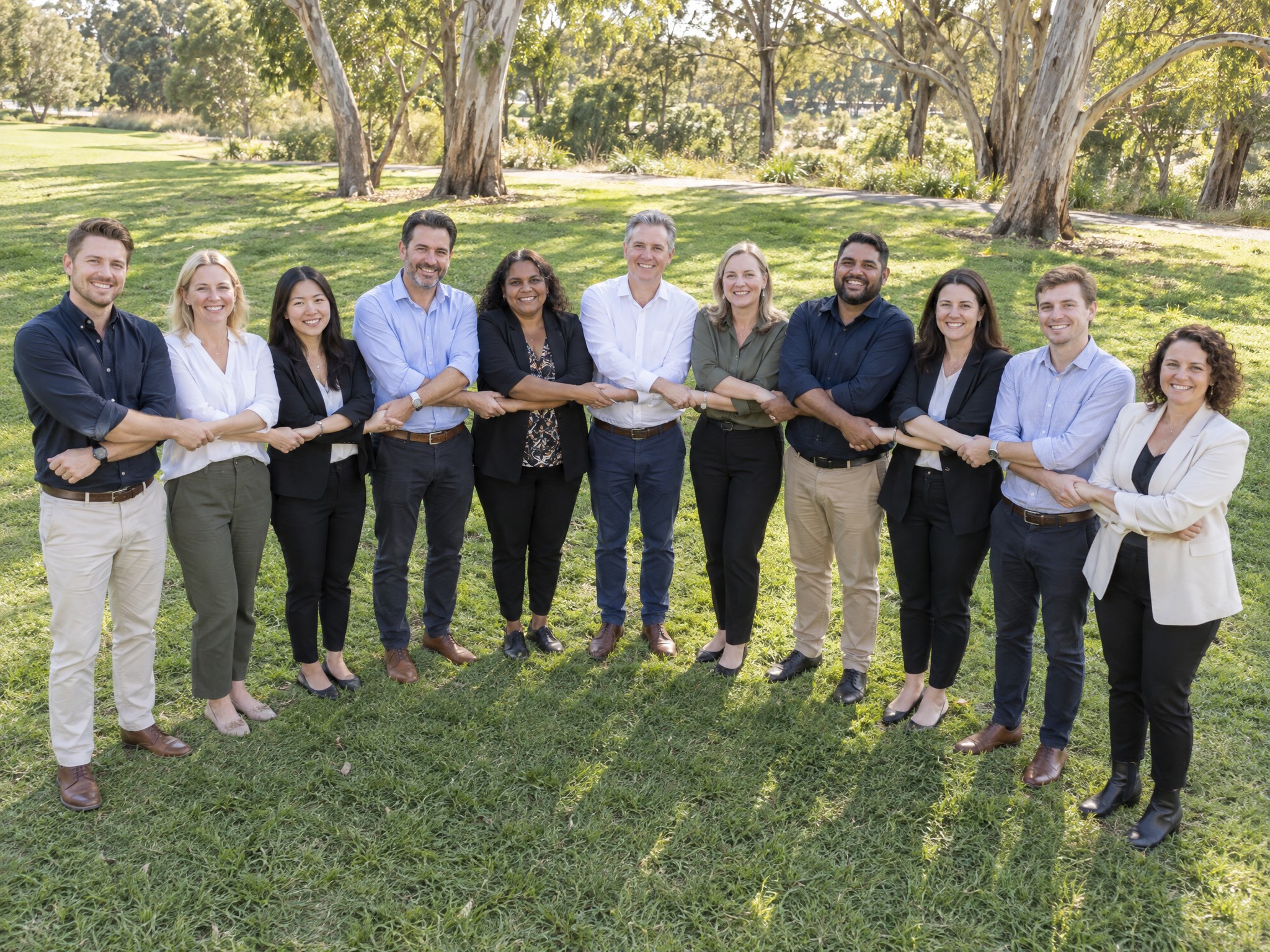 Diverse Australian colleagues laughing and holding hands in a team chain on a park lawn under gum trees